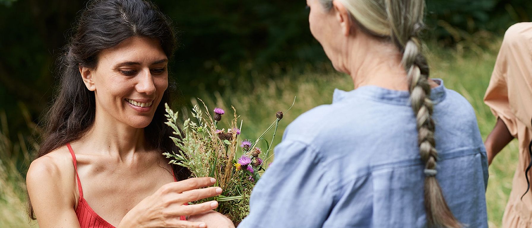 Eine juengere Frau mit dunklen langen Haaren und eine aeltere Frau mit langem grauen Zopf wenden sich einander zu, im Hintergrund eine Blumenwiese