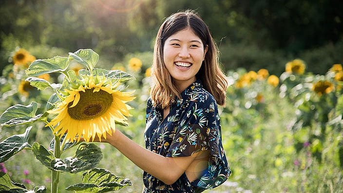 Frau steht in Sonnenblumenfeld und haelt eine Sonnenblume in der Hand