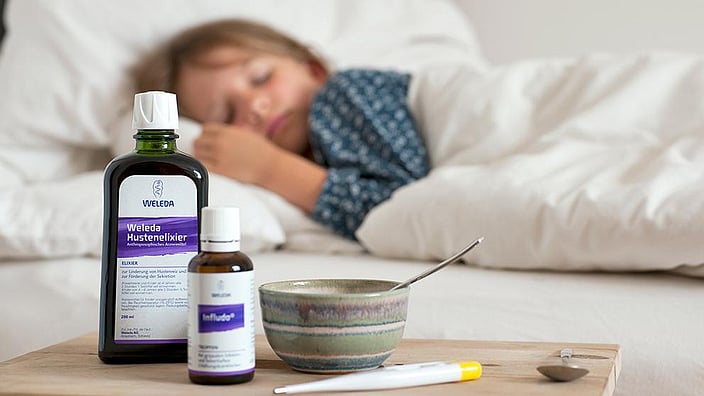 Hustenelixier, Infludo und Fiebermesser auf dem Tisch, Kind schläft im Hintergrund