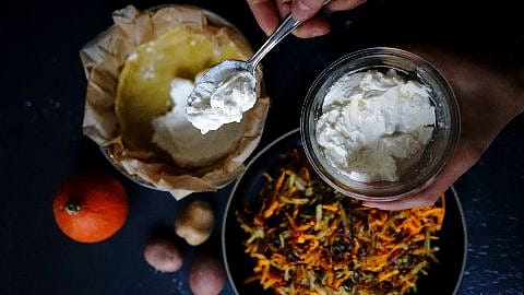 Hand haelt eine Glasschale mit Sauerrahm und einen Loeffel, darunter vorbereitete Kuchenform