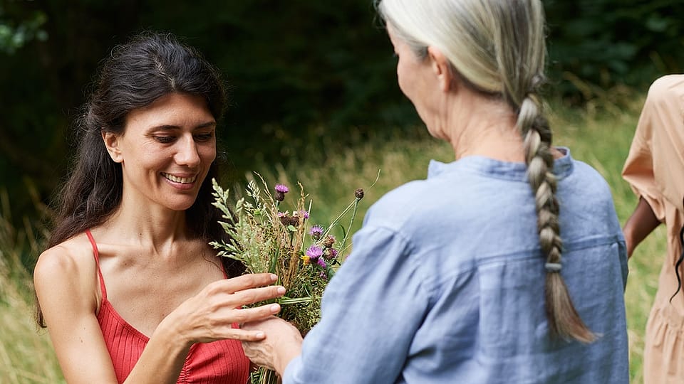 Eine juengere Frau mit dunklen langen Haaren und eine aeltere Frau mit langem grauen Zopf wenden sich einander zu, im Hintergrund eine Blumenwiese